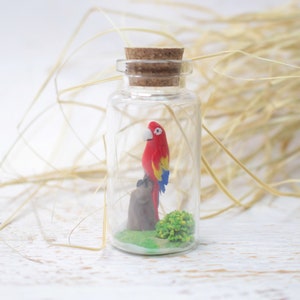 May include: A small glass bottle with a cork stopper contains a miniature red and yellow parrot perched on a small brown branch. The parrot is surrounded by green foliage.
