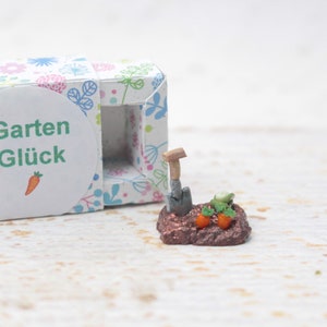 May include: A small white box with a floral pattern and the text "Garten Glück" on a white circle. The box is open and reveals a miniature garden scene with a tiny shovel, dirt, and two small carrots.
