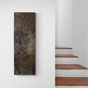 May include: A three-panel wall art piece with a wood-like texture and a wavy pattern in shades of brown and gray. The art is hanging on a white wall next to a set of wooden stairs.