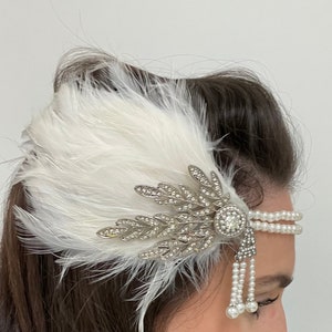 May include: A white feather headband with a silver and crystal embellishment and a pearl strand. The headband has a dangling pearl fringe.