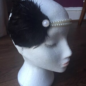 May include: A black feather headband with a pearl and rhinestone embellishment. The headband is on a white mannequin head.