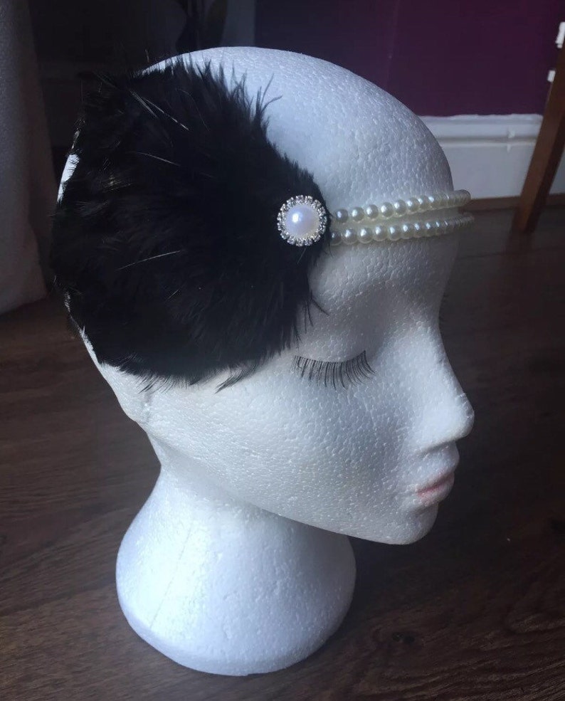 May include: A black feather headband with a pearl and rhinestone embellishment. The headband is on a white mannequin head.