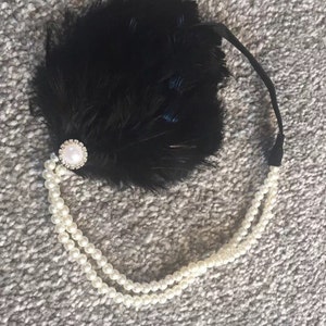 May include: A black feather headband with a pearl and rhinestone embellishment. The headband is adorned with a strand of faux pearls.