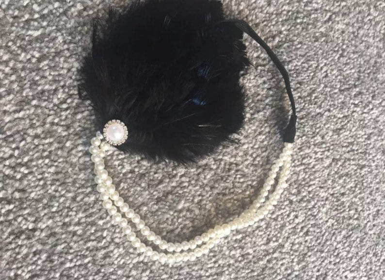 May include: A black feather headband with a pearl and rhinestone embellishment. The headband is adorned with a strand of faux pearls.