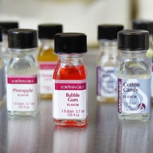 May include: A collection of small glass bottles with black lids containing different flavored extracts. The bottles are labeled with the brand name "Lorann Oils" and the flavor names, including "Pineapple", "Bubble Gum", and "Cotton Candy".