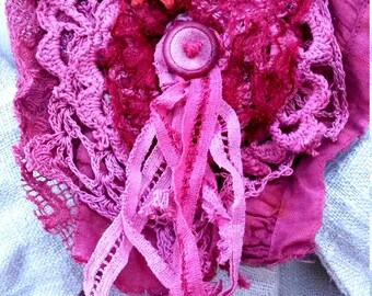 May include: A handmade, layered textile brooch in shades of pink and magenta. The design features a central button, a crocheted lace element, and flowing ribbons. A small, star-shaped embellishment adds detail. The brooch is set against a light blue fabric.