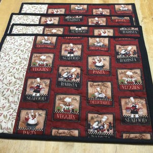 May include: A set of four quilted placemats. Each placemat features a red background with chef-themed squares, and a floral pattern on the left side. The squares have text like "Seafood" and "Pasta".
