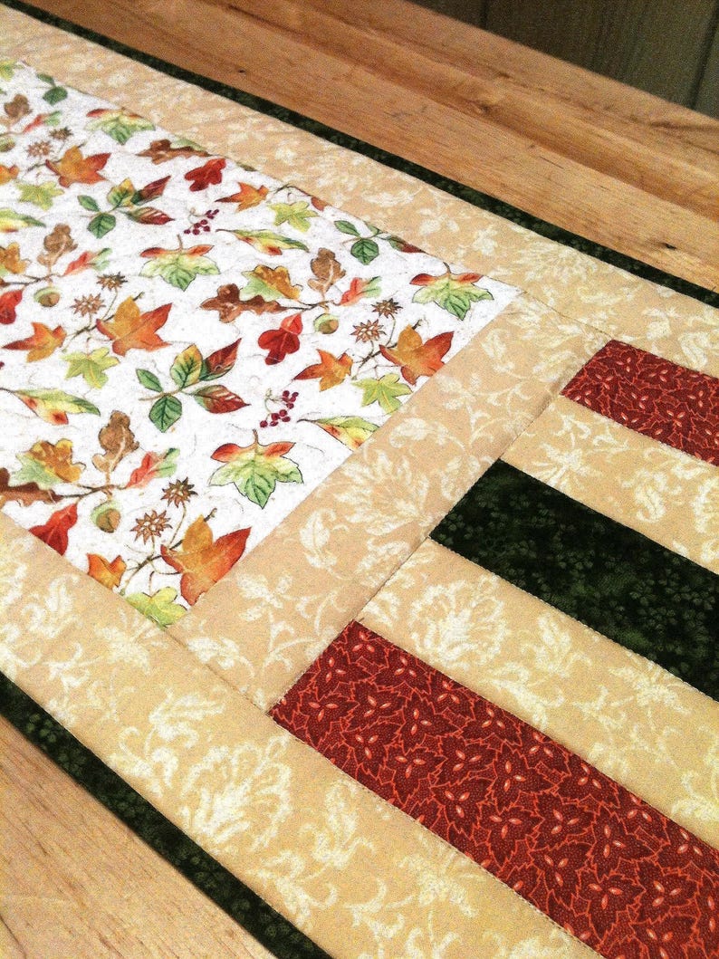 Pu&ograve; includere: Un runner da tavola trapuntato con un pannello centrale su sfondo bianco stampato con foglie autunnali colorate, ghiande e bacche. Il bordo &egrave; composto da strisce alternate di tessuto color cuoio con motivo a foglie, verde scuro e rosso.
