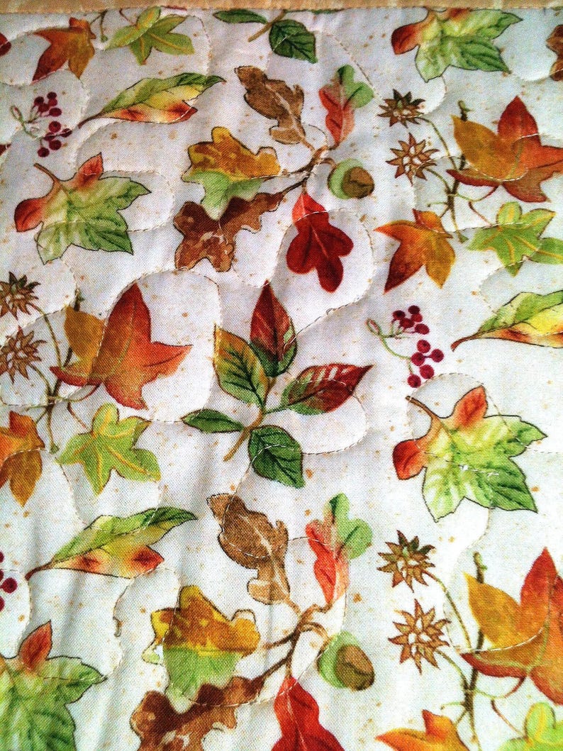 Pu&ograve; includere: Un tessuto trapuntato con sfondo color crema e un motivo ripetitivo di foglie autunnali nei toni dell'arancione, rosso, giallo e verde, intervallato da piccoli grappoli di bacche rosse e ghiande marroni, rifinito con cuciture ondulate.