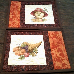May include: Two quilted placemats with a fall theme. The top placemat features a scarecrow with a red hat and a brown beard. The bottom placemat features a cornucopia with grapes, pumpkins, and an apple.