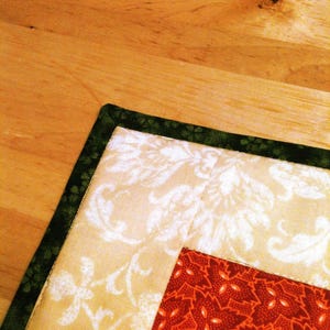 Pu&ograve; includere: Un pezzo di tessuto trapuntato caratterizzato da un bordo verde scuro con un sottile motivo floreale, una sezione color crema con una stampa damascata bianca e un blocco centrale di tessuto rosso scuro decorato con un motivo ripetuto di piccole foglie arancioni.