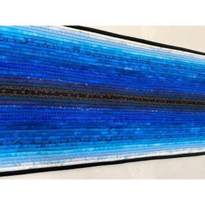 May include: A rectangular table runner featuring a gradient of blue fabric strips ranging from light sky blue to deep navy and black. The quilted textile displays various patterns including geometric shapes, dots, and floral motifs with black binding.
