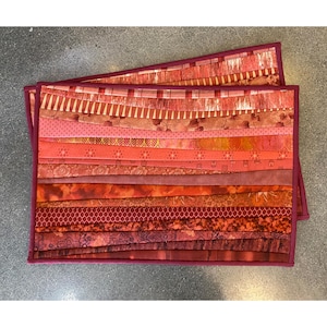 May include: A rectangular placemat constructed from horizontal fabric strips in various shades of red, orange, and burgundy. The strips feature diverse patterns including floral, geometric, and textured designs, all enclosed within a solid dark red border.