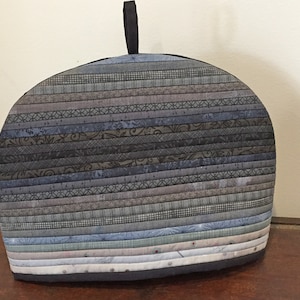 May include: A quilted tea cozy in shades of blue, gray, and brown, with a black handle. The cozy is made of horizontal strips of fabric, creating a striped pattern. The cozy is approximately 10 inches wide and 8 inches tall.