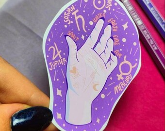 Puede incluir: Pegatina morada con una ilustración de lectura de la palma de la mano. La mano está rodeada de estrellas y planetas con las palabras "Saturno", "Espíritu", "Agua", "Tierra", "Fuego", "Aire", "Sol", "Mercurio" y "Júpiter 24".