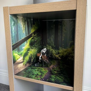May include: A wooden display case with a forest backdrop featuring a Star Wars scene. The case has a light wood frame and a detailed forest scene printed on the back. A Stormtrooper figure is mounted on a speeder bike.