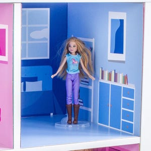 May include: A dollhouse room with blue walls, a bed, a bookshelf, and a doll wearing a blue shirt, purple trousers, and brown boots. The doll is standing on a clear plastic stand.