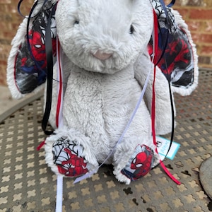May include: A plush gray rabbit stuffed animal featuring long ears lined with a fabric pattern of Spider-Man and spiderwebs. The rabbit is adorned with multiple thin satin ribbons in navy blue, black, red, and white, and has matching fabric on its feet.