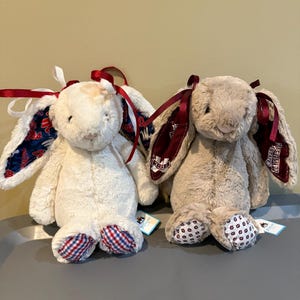 May include: Two plush stuffed rabbits with long ears. The left rabbit is cream-colored with blue and red patterned ear linings and red and white ribbons. The right rabbit is tan with maroon Mississippi State branded ear linings and maroon ribbons.