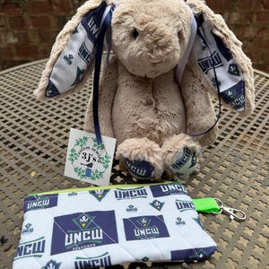 May include: A plush tan bunny with blue ribbon accents and patterned ears, feet, and a matching zippered pouch. The pouch and ear lining feature a white background with navy blue and green text and logos. A tag reads "custom sewing 3j's".