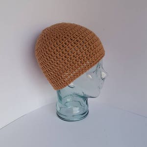 May include: A tan crochet beanie hat displayed on a glass head. The hat has a textured, open weave design. The hat is a neutral color and appears to be handmade. The background is white.