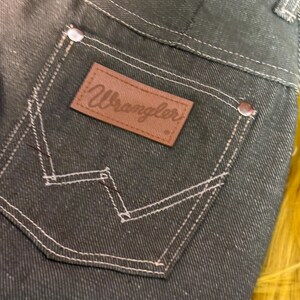 May include: Close-up of dark denim jeans with a Wrangler brand patch on the back pocket. The jeans feature white stitching and metal rivets. The denim has a speckled appearance.