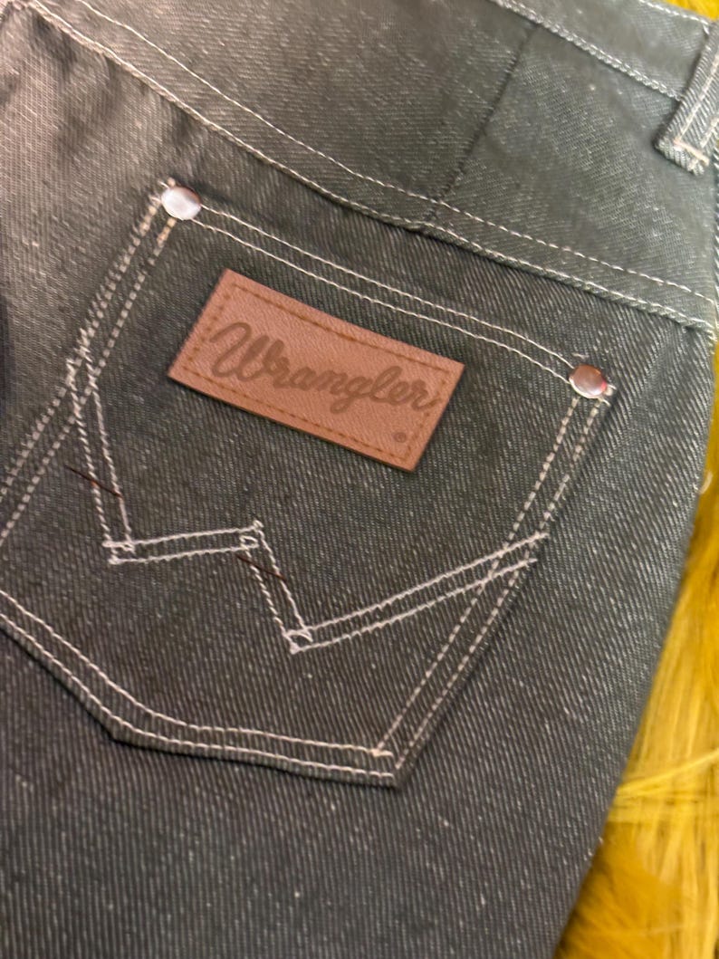 May include: Close-up of dark denim jeans with a Wrangler brand patch on the back pocket. The jeans feature white stitching and metal rivets. The denim has a speckled appearance.