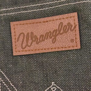 May include: Close-up of a brown leather Wrangler brand patch sewn onto dark green denim fabric. The patch has the Wrangler logo in a cursive font. White stitching is visible around the patch and on the denim.