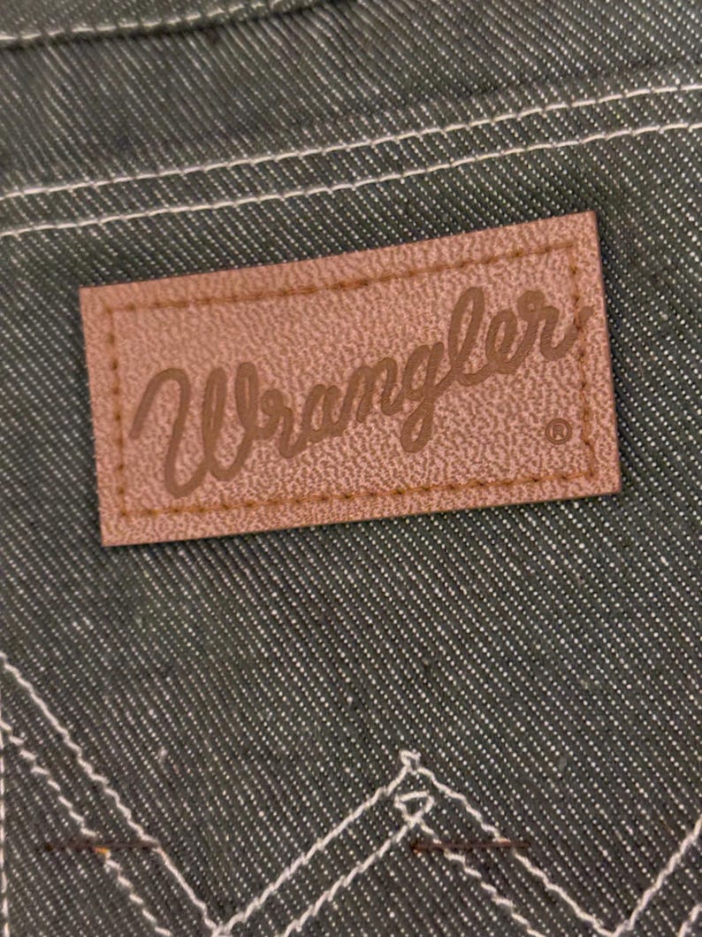 May include: Close-up of a brown leather Wrangler brand patch sewn onto dark green denim fabric. The patch has the Wrangler logo in a cursive font. White stitching is visible around the patch and on the denim.
