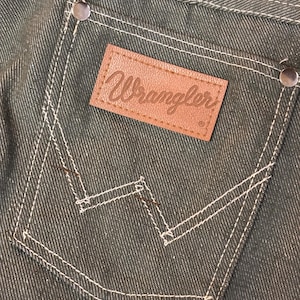 May include: Close-up of a dark green denim pocket with white stitching and a brown leather Wrangler label. The pocket has a classic design with a signature "W" stitch detail and metal rivets.