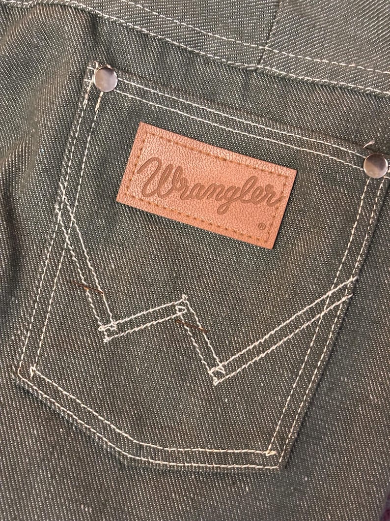 May include: Close-up of a dark green denim pocket with white stitching and a brown leather Wrangler label. The pocket has a classic design with a signature "W" stitch detail and metal rivets.