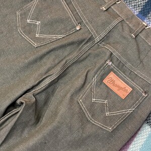 May include: Close-up of the back of dark green Wrangler jeans. The jeans have white stitching, two back pockets with a signature "W" design, and a brown leather patch with the Wrangler logo. The jeans are made of a durable denim material.
