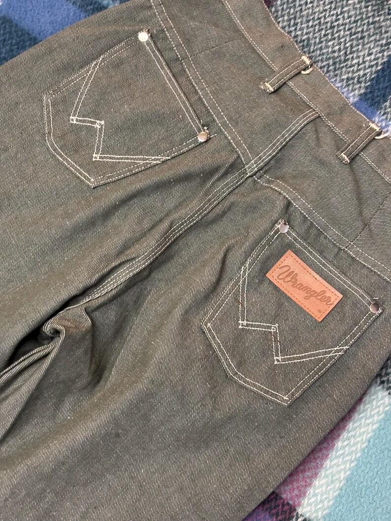 May include: Close-up of the back of dark green Wrangler jeans. The jeans have white stitching, two back pockets with a signature "W" design, and a brown leather patch with the Wrangler logo. The jeans are made of a durable denim material.