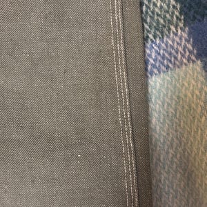 May include: Close-up of gray denim fabric with white stitching along the seam. The fabric has a textured appearance. The edge of a blue, green, and white plaid blanket is visible in the background.