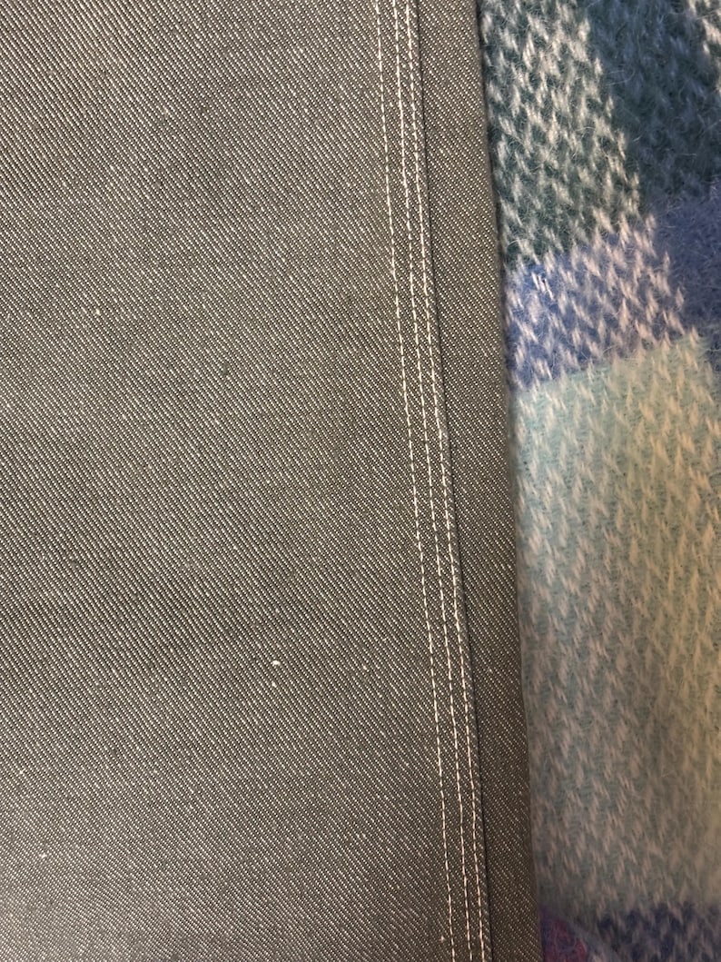May include: Close-up of gray denim fabric with white stitching along the seam. The fabric has a textured appearance. The edge of a blue, green, and white plaid blanket is visible in the background.