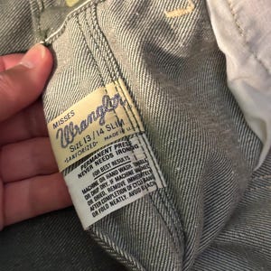 May include: Close-up of a pair of gray Wrangler jeans. The label reads "MISSES Wrangler SIZE 13/14 SLIM SANFORIZED MADE IN U.S.A." Additional text on the label provides care instructions. The jeans are made of a durable denim fabric.