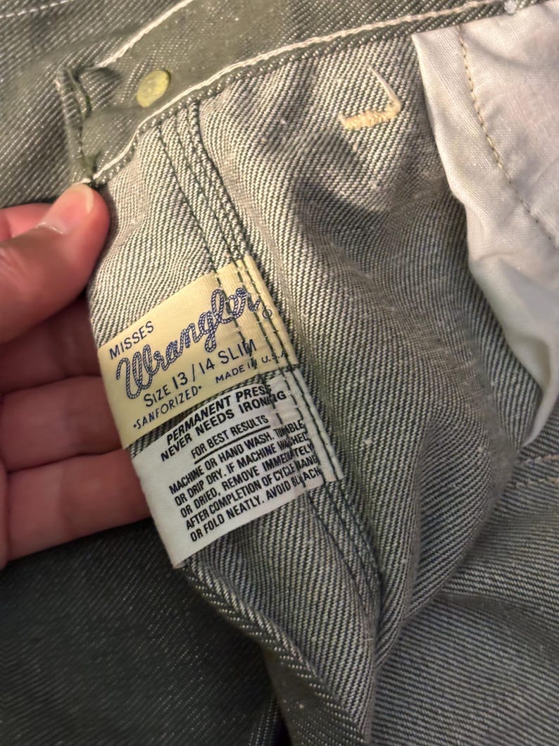 May include: Close-up of a pair of gray Wrangler jeans. The label reads "MISSES Wrangler SIZE 13/14 SLIM SANFORIZED MADE IN U.S.A." Additional text on the label provides care instructions. The jeans are made of a durable denim fabric.