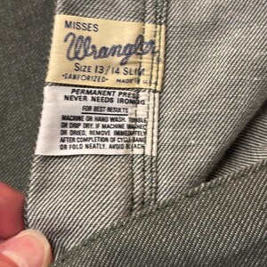 May include: Close-up of a pair of gray Wrangler jeans. The label reads "MISSES Wrangler SIZE 13/14 SLIM SANFORIZED MADE IN U.S.A." with care instructions. The jeans are made of a durable denim fabric.