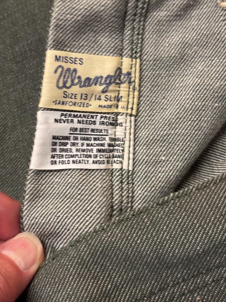 May include: Close-up of a pair of gray Wrangler jeans. The label reads "MISSES Wrangler SIZE 13/14 SLIM SANFORIZED MADE IN U.S.A." with care instructions. The jeans are made of a durable denim fabric.