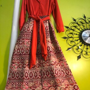 May include: A long, orange dress with a patterned skirt. The skirt has a geometric pattern in shades of red, pink, and white. The dress has a long sleeve and a tie at the waist.