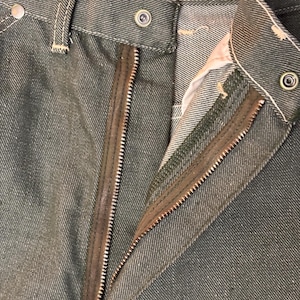 May include: Close-up of olive green denim pants with a partially unzipped zipper. The pants have a button closure and white stitching details. The zipper is brown and the fabric has a textured appearance.