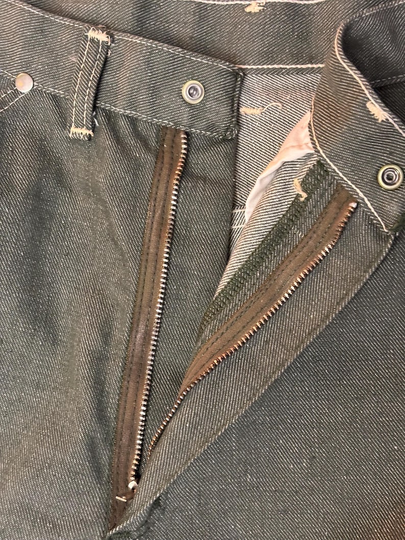 May include: Close-up of olive green denim pants with a partially unzipped zipper. The pants have a button closure and white stitching details. The zipper is brown and the fabric has a textured appearance.