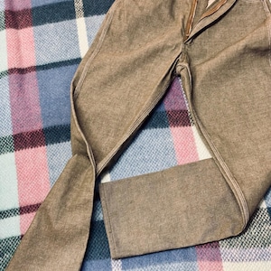 May include: Brown denim pants with a zipper fly and button closure. The pants have white stitching along the seams and pockets. The pants are folded on a plaid blanket.