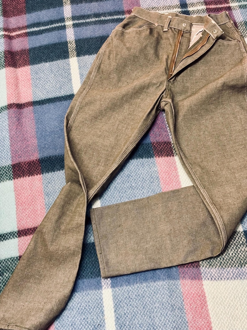 May include: Brown denim pants with a zipper fly and button closure. The pants have white stitching along the seams and pockets. The pants are folded on a plaid blanket.