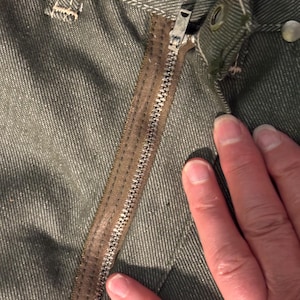 May include: Close-up of olive green denim pants with a brown zipper and metal button details. The zipper is partially open, revealing the metal teeth. The fabric has a textured appearance.