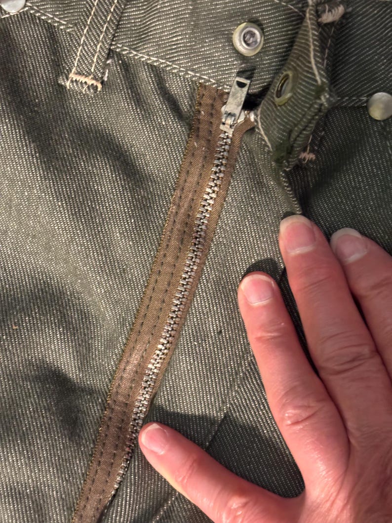 May include: Close-up of olive green denim pants with a brown zipper and metal button details. The zipper is partially open, revealing the metal teeth. The fabric has a textured appearance.