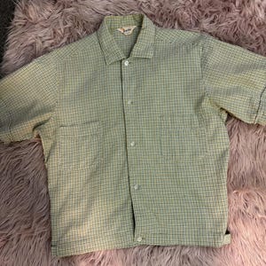 May include: A light green and white checkered button-up shirt with short sleeves. The shirt has two front pockets and a collar. The brand name "Bogie" is visible on the label. The shirt is laid flat on a pink, fluffy surface.