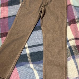 May include: Brown denim pants with visible white stitching. The pants have a classic five-pocket design and a button and zipper closure. The pants are made of a sturdy material and appear to be new.
