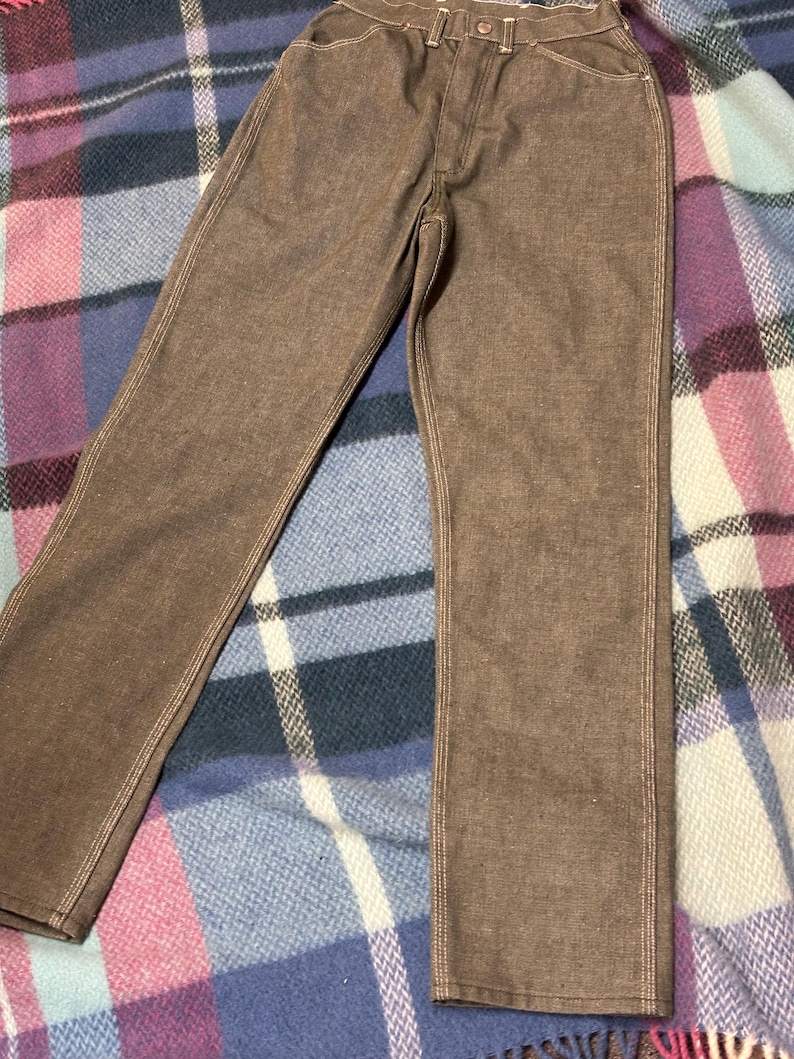 May include: Brown denim pants with visible white stitching. The pants have a classic five-pocket design and a button and zipper closure. The pants are made of a sturdy material and appear to be new.