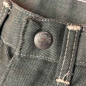 May include: Close-up of denim pants with a silver button and white stitching. The button has an embossed design. The fabric is a dark gray-green color. The stitching is visible along the seams and around the button.